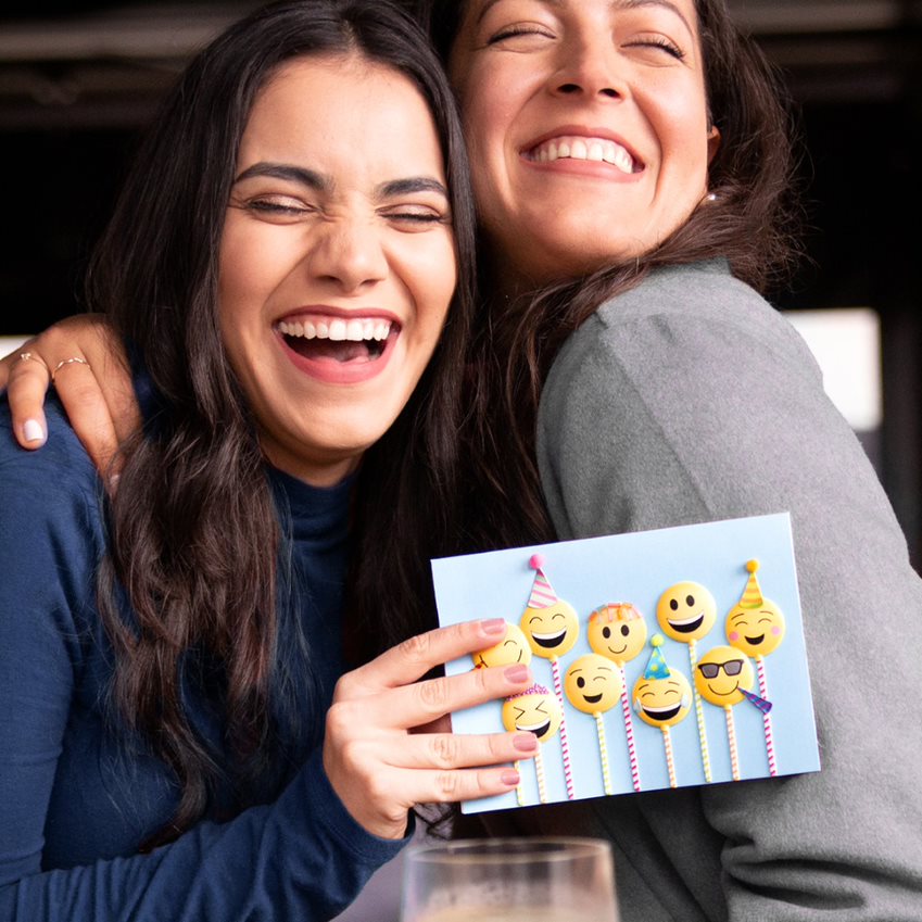 Two friends hugging, laughing, and holding an emoji birthday greeting card